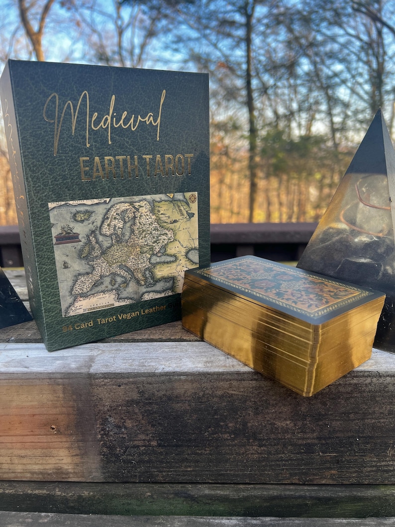 May include: A Medieval Earth Tarot deck in a green box with gold lettering and a map design. The deck is next to a box of cards with gold edges. The box reads "54 Card Tarot Vegan Leather".