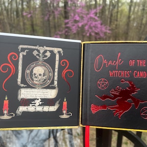 May include: A black book titled "Oracle of the Witches' Candle" with red lettering and a witch graphic. The open book reveals a skull illustration, candles, and scrollwork. The book has a gold trim.