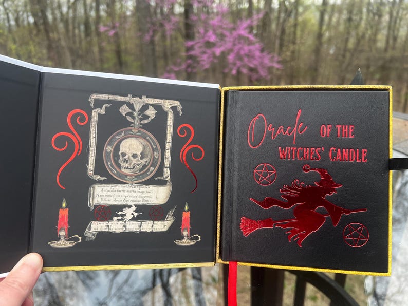 May include: A black book titled "Oracle of the Witches' Candle" with red lettering and a witch graphic. The open book reveals a skull illustration, candles, and scrollwork. The book has a gold trim.