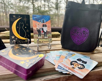 May include: A deck of tarot cards with black cats and a crescent moon design. The cards are displayed on a wooden table with a black leather pouch with a purple Celtic knot heart design. The cards are labeled with the names of the suits, such as "Ace of Cups".