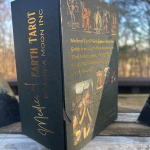 May include: A black tarot card box with gold lettering that reads "Medieval Earth Tarot by Libra Moon Inc." The box features medieval-style illustrations and text. The color scheme is primarily black, green, and gold.