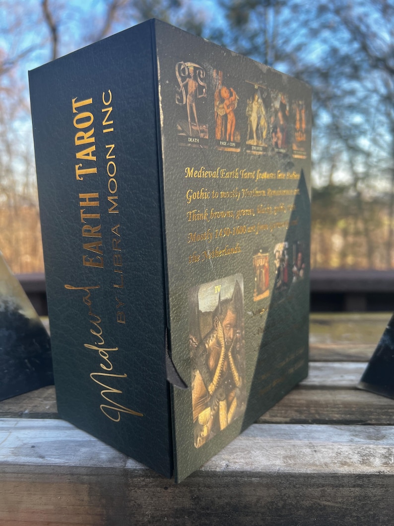 May include: A black tarot card box with gold lettering that reads "Medieval Earth Tarot by Libra Moon Inc." The box features medieval-style illustrations and text. The color scheme is primarily black, green, and gold.