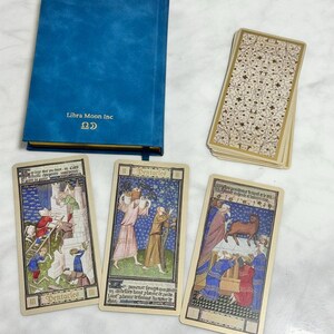 May include: A blue hardcover book with the text Libra Moon Inc and a symbol on the cover, accompanied by a deck of cards with a cream and gold floral pattern and three tarot cards featuring medieval illustrations and the word Pentacles.
