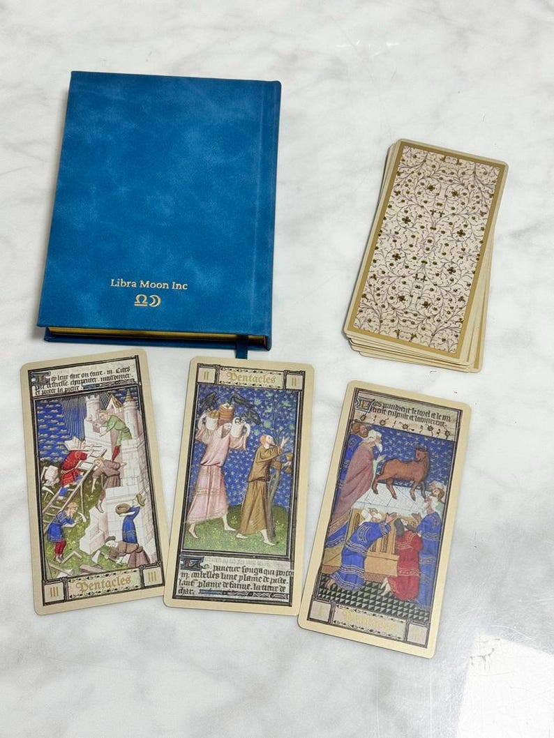 May include: A blue hardcover book with the text Libra Moon Inc and a symbol on the cover, accompanied by a deck of cards with a cream and gold floral pattern and three tarot cards featuring medieval illustrations and the word Pentacles.