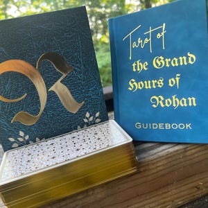 Könnte beinhalten: Ein Tarotkartendeck mit goldfarbenen Kanten und gemusterten Rückseiten, präsentiert neben einer dunkel-türkisen Schachtel mit einem großen goldenen Buchstaben R und einem blauen Begleitbuch mit dem Titel Tarot of the Grand Hours of Rohan.