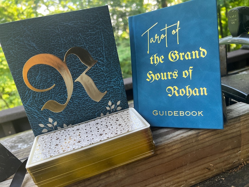 May include: A deck of tarot cards with gold-gilded edges and a patterned back, displayed alongside a dark teal box featuring a large gold letter R and a blue guidebook titled Tarot of the Grand Hours of Rohan in gold lettering.