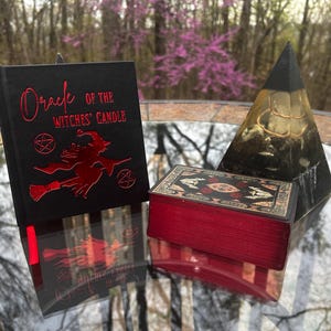 May include: A black book with the title "Oracle of the Witches' Candle" in red lettering, featuring a witch on a broom. A red-edged tarot card box and a black and gold pyramid are also present.