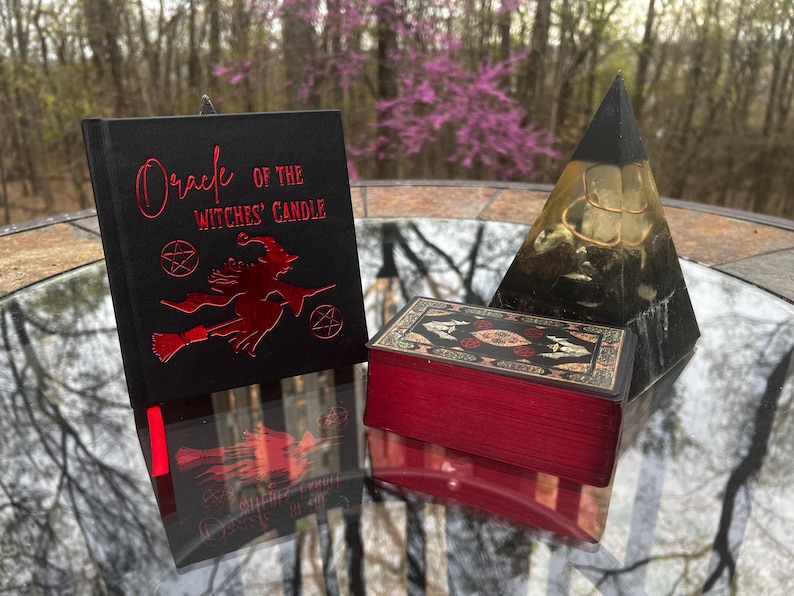 May include: A black book with the title "Oracle of the Witches' Candle" in red lettering, featuring a witch on a broom. A red-edged tarot card box and a black and gold pyramid are also present.