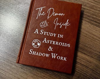 Może przedstawiać: Brązowa książka w twardej oprawie z białym napisem The Demon Inside: A Study in Asteroids & Shadow Work oraz dwiema małymi białymi ilustracjami liniowymi przedstawiającymi asteroidy obok tytułu i podtytułu.