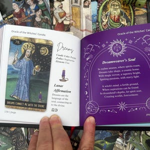 May include: An open book displaying two pages from an oracle card deck. One page features a card with a figure in blue robes, the text "Dreams Connect Me With The Divine", and the number 78. The opposite page has purple text and illustrations.