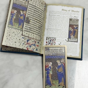 May include: An open book with medieval-style illuminated manuscript pages featuring Latin text and colorful illustrations of figures in robes, alongside a matching tarot card titled King of Wands, which depicts a crowned figure in a blue cloak.
