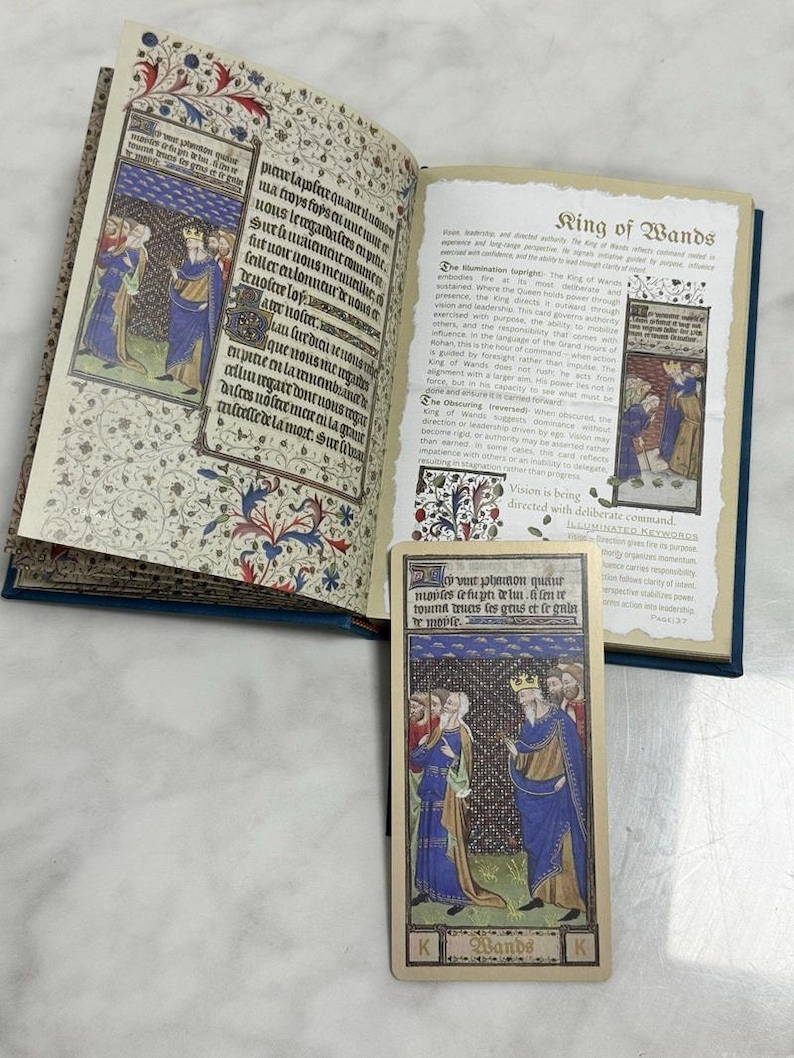 May include: An open book with medieval-style illuminated manuscript pages featuring Latin text and colorful illustrations of figures in robes, alongside a matching tarot card titled King of Wands, which depicts a crowned figure in a blue cloak.