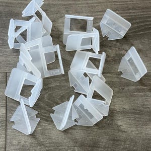 May include: A collection of translucent white plastic tile leveling spacers. Each spacer has a rectangular opening and the text "3.0 mm" is visible on some. The spacers are scattered on a wooden surface.