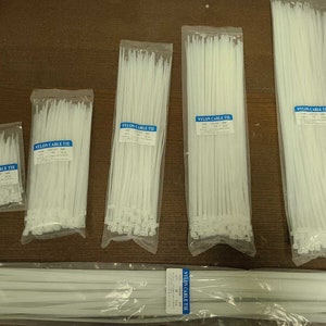 May include: Several clear plastic bags filled with white nylon cable ties are arranged on a brown surface. Each bag has a blue label that reads "NYLON CABLE TIE". The bags vary in size, with the largest containing a long bundle of ties.