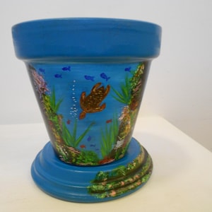 May include: A blue ceramic flower pot featuring a hand-painted underwater scene. The design includes a brown sea turtle swimming among green aquatic plants, coral formations, and small blue fish, with bubbles rising toward the top rim.