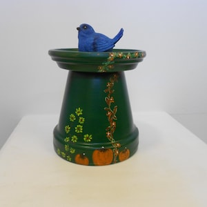 May include: A dark green painted terracotta pot with a wide rim, featuring hand-painted orange pumpkins and green floral vines. A small, bright blue bird figurine is perched on the flat top surface of the inverted pot structure.