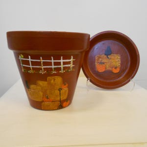 May include: A terracotta flower pot and matching saucer featuring hand-painted autumn scenes. The pot displays a white fence, hay bales, pumpkins, and a black cat. The saucer shows a black cat sitting on a hay bale with two orange pumpkins.