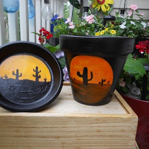 May include: A black terracotta plant pot and matching saucer, both hand-painted with a desert landscape featuring a bright orange sun and black silhouettes of saguaro cacti. The exterior of the pot and the interior of the saucer display this scene.