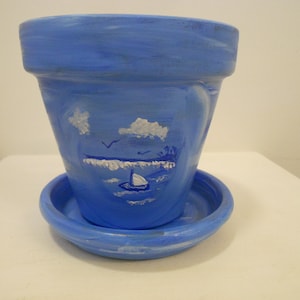 May include: A blue ceramic flower pot with a matching saucer, featuring a hand-painted white scene of a sailboat on the water, clouds in the sky, and birds flying above. The pot has a wide rim and a tapered body with visible brush strokes.