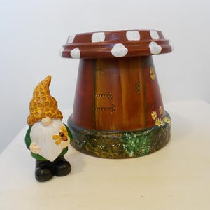 May include: A small gnome figurine with a yellow honeycomb-patterned hat and a white beard holding a bee, positioned next to a brown, inverted terracotta pot painted to resemble a mushroom house with a door, windows, and floral details.