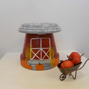 May include: A red painted terracotta pot shaped like a barn with a gray lid, featuring white window frames, yellow corn stalks, and orange pumpkins. Beside it sits a small metal wheelbarrow containing two orange pumpkins.