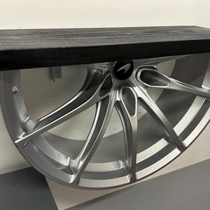 May include: A silver car wheel with a black wooden shelf attached to it. The wheel has a silver finish and a black logo on the side. The shelf is made of wood and has a black finish.