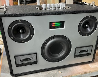Op de afbeelding: Een zilveren en zwarte boombox met een grote luidspreker in het midden, twee kleinere luidsprekers aan de zijkanten en een cassettedeck aan de rechterkant. De boombox heeft een groen en rood LED-display en vijf knoppen aan de bovenkant.