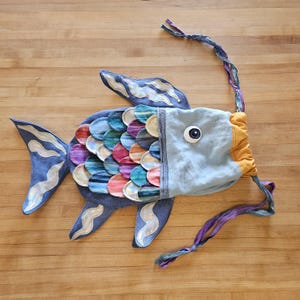 Op de afbeelding: Een grillige visvormige tas met een blauwe denim body en kleurrijke, gelaagde schubben. De tas heeft een lichtblauwe kop met een groot oog en een oranje trekkoordsluiting. Een lange, gevlochten band voegt een speels tintje toe.