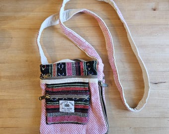 May include: A pink and white crossbody bag with a patterned front pocket and a long strap. The bag has a zipper closure and a small tag that reads "NATURAL HEMP 100% HEMP". The bag is made of a woven fabric.