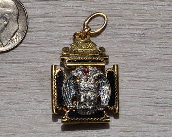 May include: A gold-colored pendant with a black and white eagle design. The eagle has a red jewel in its chest. The pendant is surrounded by a gold frame with a decorative top.