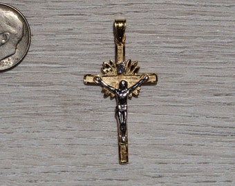 Op de afbeelding: Een goudkleurige kruisbeeldhanger met een zilverkleurige figuur van Jezus. Het kruis heeft een zonnestraalontwerp achter de figuur. Een Amerikaanse dime ligt ernaast ter vergelijking.