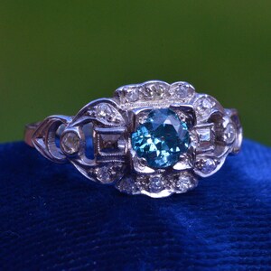 K&ouml;nnte beinhalten: Ein silberner Ring mit einem blauen Edelstein, der von kleineren Diamanten umgeben ist. Der Ring hat ein zartes, filigranes Design.
