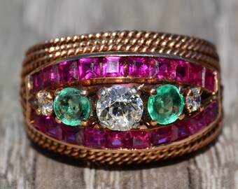 Puede incluir: Un anillo de oro con un gran diamante redondo y claro rodeado de dos esmeraldas verdes y diamantes más pequeños. El anillo está engastado con una banda de rubíes rosas.