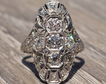 May include: A silver ring with a large oval-shaped setting featuring a cluster of diamonds. The ring has a delicate filigree design and is set with multiple smaller diamonds.