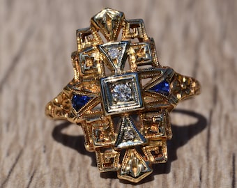 Può includere: Un anello d'oro con un design geometrico che presenta un diamante quadrato centrale e tre zaffiri blu. L'anello ha un motivo a filigrana dettagliato.