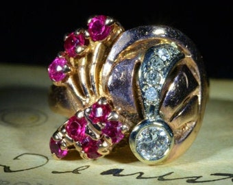 Può includere: Un anello d'oro con un grande diamante e un gruppo di rubini. L'anello è incastonato in un design unico con una fascia curva e una montatura rialzata per il diamante.