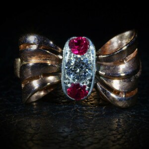 Op de afbeelding: Een gouden ring met een strikdesign met een grote diamant en twee robijnen.