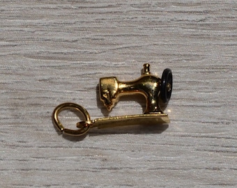 May include: A gold-colored sewing machine charm with a black wheel, next to a U.S. dime. The charm has a small loop for attaching to a bracelet or necklace. The dime is silver and shows a profile.