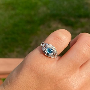 K&ouml;nnte beinhalten: Ein silberner Ring mit einem blauen Edelstein, der von kleineren, klaren Edelsteinen umgeben ist.