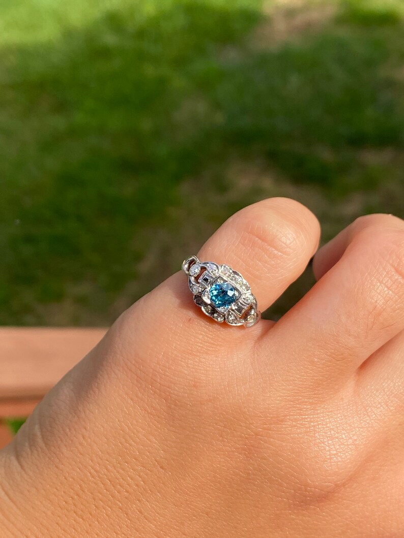 K&ouml;nnte beinhalten: Ein silberner Ring mit einem blauen Edelstein, der von kleineren, klaren Edelsteinen umgeben ist.