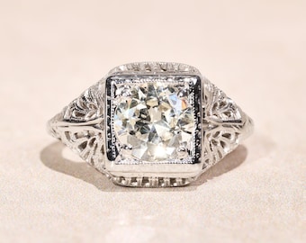 Könnte beinhalten: Ein silberfarbener Ring mit einem runden, facettierten, klaren Edelstein in einer quadratischen Fassung. Die Ringschiene und die Fassung weisen filigrane, durchbrochene Metallverzierungen auf, die sich entlang der Seiten des Rings erstrecken.