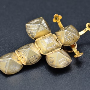 May include: A gold-toned, hinged, cross-shaped pendant with intricate geometric designs on each side. The pendant is made of multiple interlocking pieces.