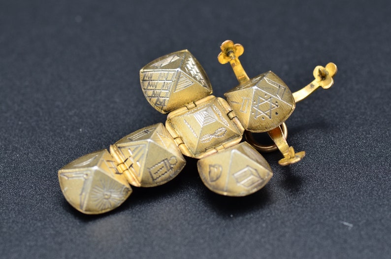 May include: A gold-toned, hinged, cross-shaped pendant with intricate geometric designs on each side. The pendant is made of multiple interlocking pieces.