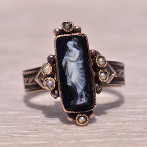 Pu&ograve; includere: Anello antico con cammeo nero rettangolare raffigurante una figura bianca. La fascia color oro ha incisioni decorative ed &egrave; impreziosita da piccole perle. Un gioiello classico.