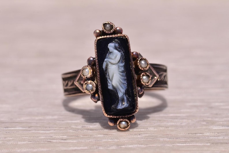 Pu&ograve; includere: Anello antico con cammeo nero rettangolare raffigurante una figura bianca. La fascia color oro ha incisioni decorative ed &egrave; impreziosita da piccole perle. Un gioiello classico.