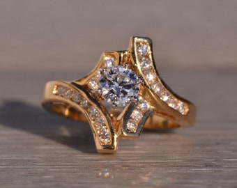 May include: A gold ring with a large, clear, round diamond in the center. The ring is set with smaller diamonds around the center stone and on the band.