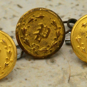 Könnte beinhalten: Drei goldfarbene Knöpfe mit einem erhabenen Design aus einem Kranz und einem chinesischen Schriftzeichen. Die Knöpfe befinden sich auf einer hellbraunen Oberfläche.