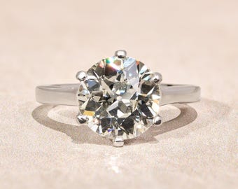 May include: A silver-coloured metal ring featuring a large, round, faceted clear stone set in a six-prong setting. The band is smooth and polished, with the central stone displaying multiple reflective facets that catch the light.