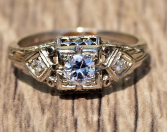 Pode incluir: Um anel de noivado vintage com um diamante central rodeado por diamantes menores. O anel é engastado em metal branco com detalhes intrincados de filigrana.