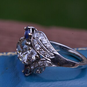 May include: A silver ring featuring a large, clear, round diamond surrounded by smaller diamonds and two blue sapphires.